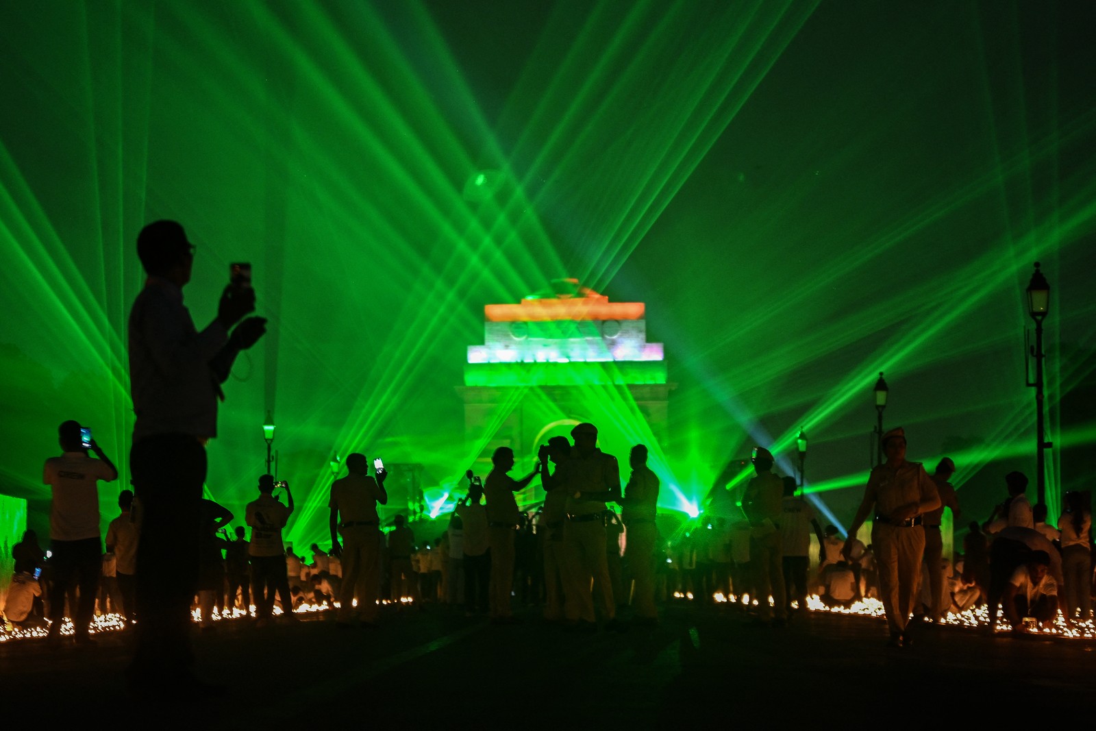 Many people stand around, taking photographs and video of lamps arranged in a large square, beneath a laser light show.