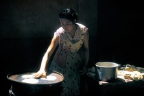 A woman lit by a ray of light against a black background lays out tortillas she's cut from dough