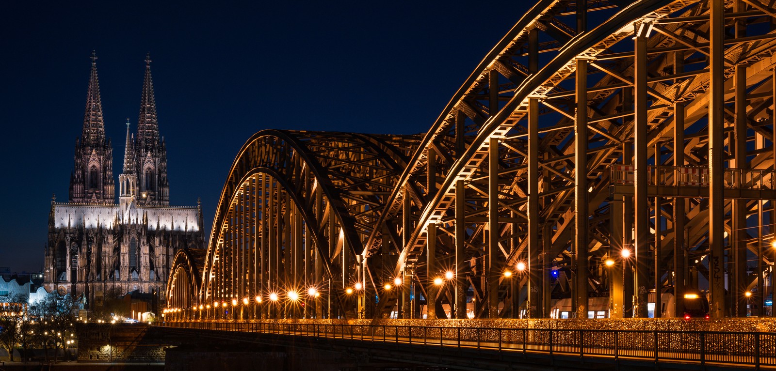 A night view of an illuminated rail bridge and tall cathedral in Germany.
