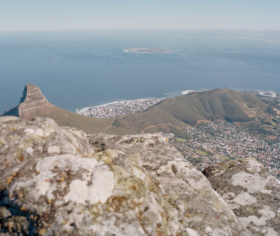 photograph looking down from rocky outcrop at distant ridge of mountain, city both in foreground and on other side, with shore and ocean beyond and small island visible