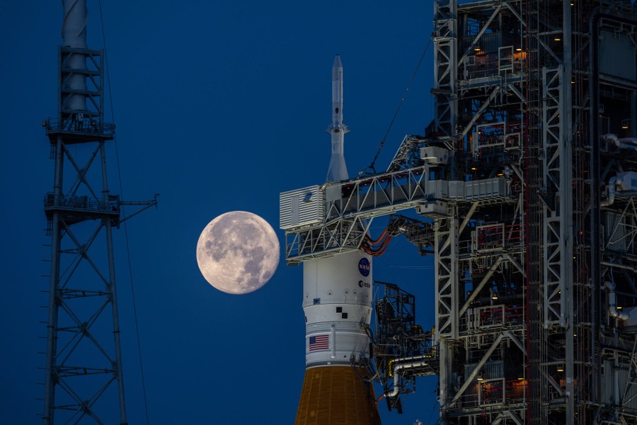 The full moon is seen in the sky behind the top section of a large rocket on a launch pad.