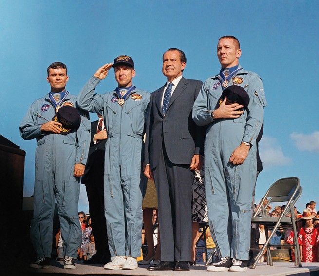 color photo of Nixon in suit and tie standing on outdoor stage next to 3 astronauts in blue coveralls, two holding hats to their hearts and one saluting, with crowd of onlookers and blue sky in background