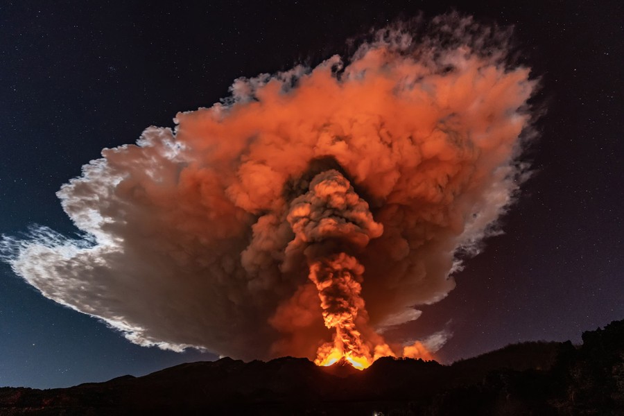 Reddish light from lava illuminates the base of a column of ash rising into a cloud above a volcano.