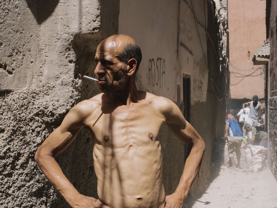 A man stands in an alley, smoking a cigarette, as people behind him work to clear rubble.