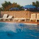 A slightly fuzzy, film-style photograph of a pool lined with white pool chairs and blue pool umbrellas, with water splashing from the center.