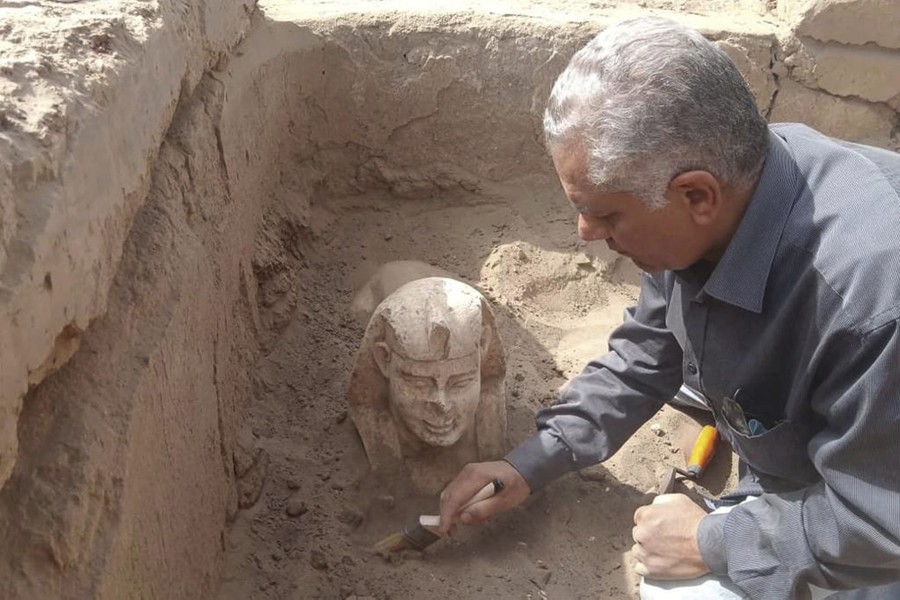 A person in a shallow pit uses a brush to help expose a small buried sphinx statue.