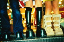 Three people are wearing boots while standing on an escalator. The image is taken from the thigh down, with a mall in the background