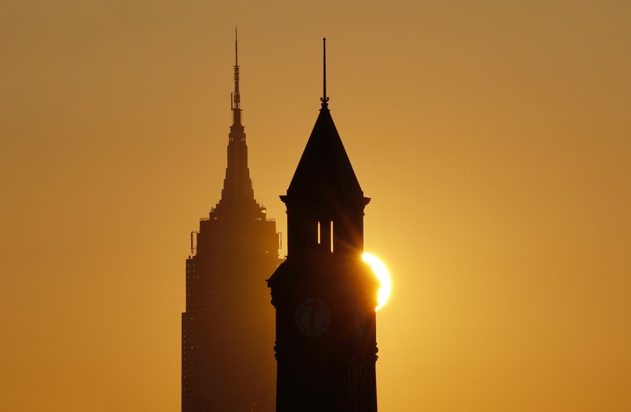 The sun rises behind two tall buildings.