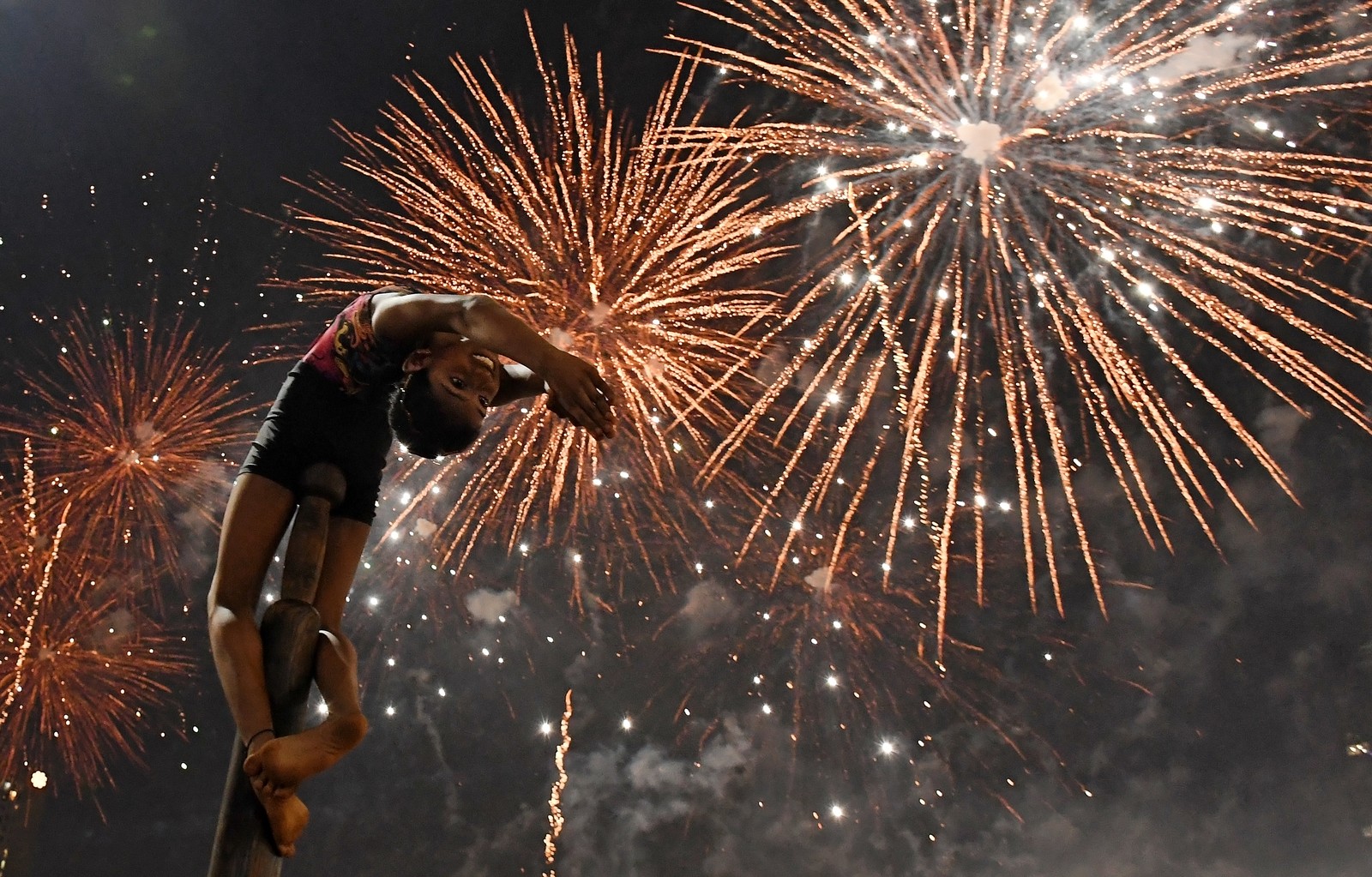 A girl performs gymnastics on a pole, as fireworks light up the sky above.