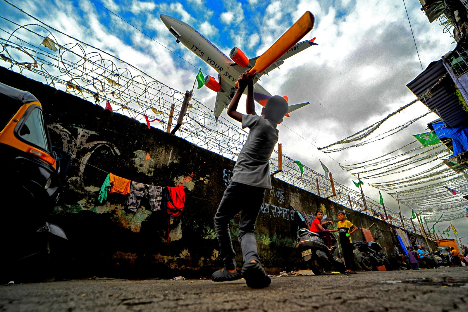 Children play a game of cricket in a road beside a long fence, as an aircraft passes close overhead.