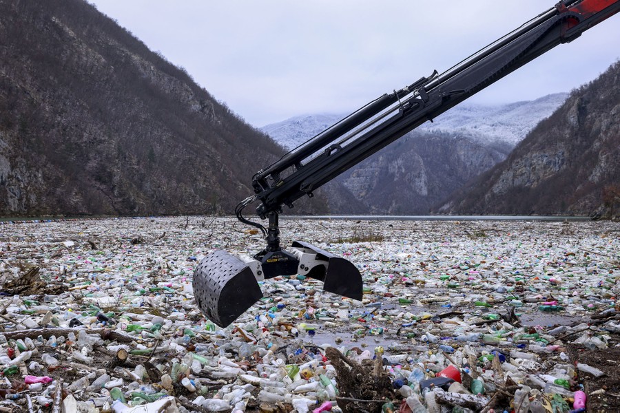 A crane holds an open bucket above a large patch of floating garbage on a river.