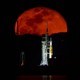 A rocket stands on a launch pad, lit at night, seen from a distance, with a red-colored full moon rising in the background.
