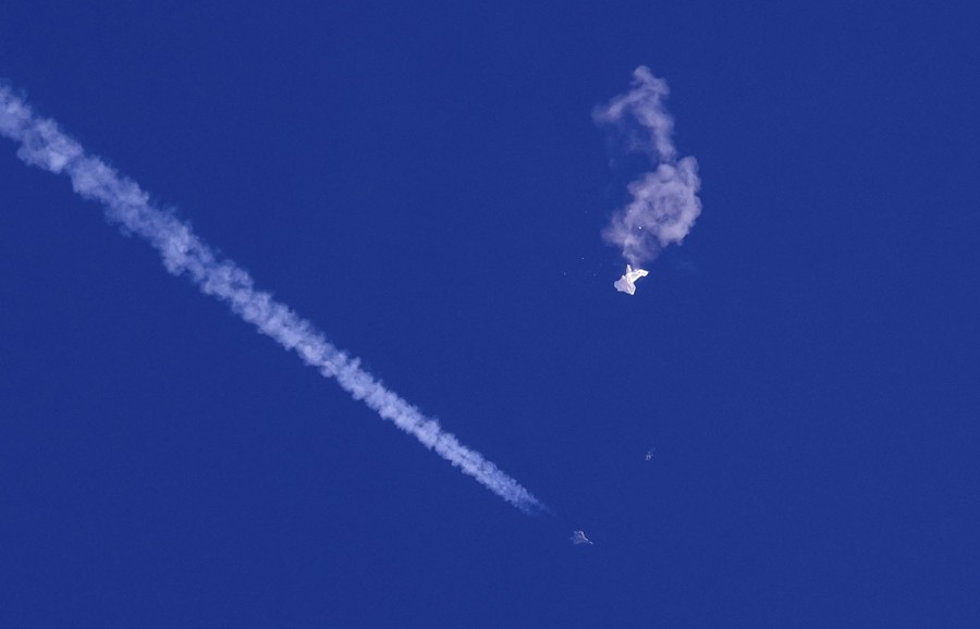 A fighter jet flies near the remnants of a large balloon that has burst and is falling.