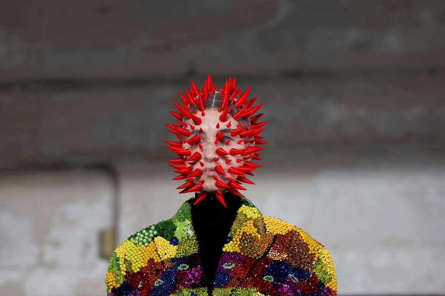 A model wears a colorful jacket and a clear mesh mask covered with red spikes.