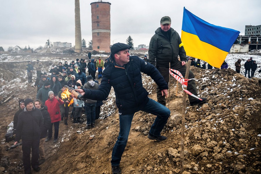 Several dozen people stand together along a small dirt hill in a vacant lot, waiting behind a person who is throwing a Molotov cocktail, watched by a trainer.