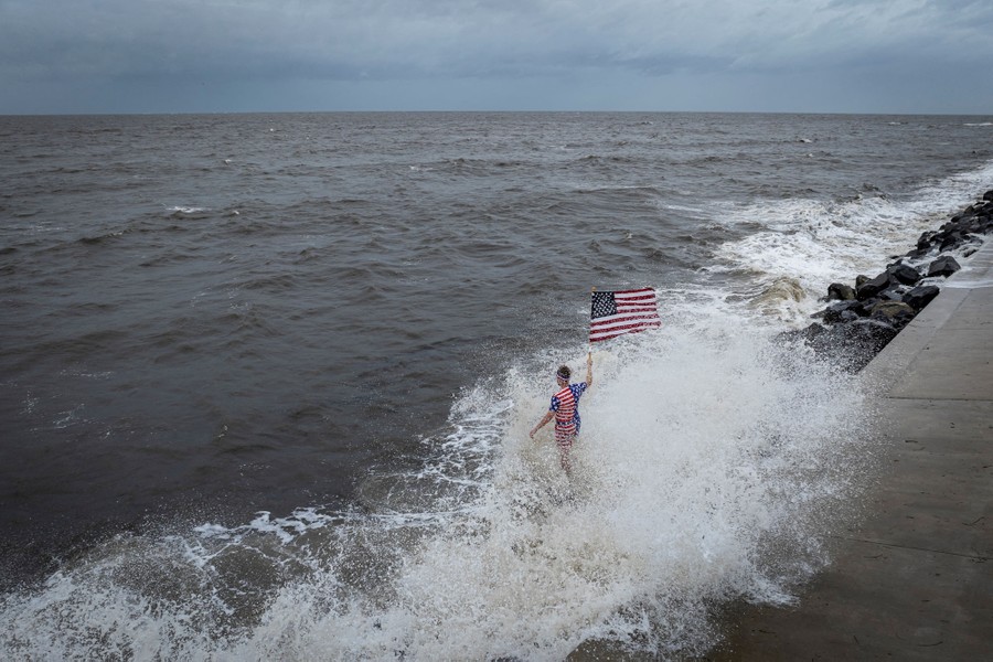 A person wearing American flag-themed shorts and shirt waves an American flag while standing in crashing surf, facing out toward dark clouds on the horizon.