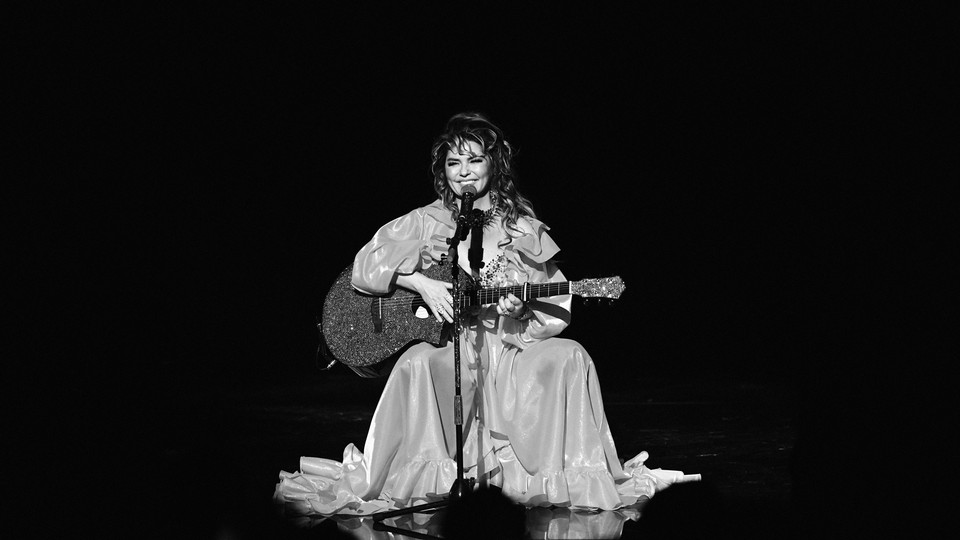 A black-and-white photo of Shania Twain performing onstage. She's seated behind a microphone, wearing a voluminous, frilly dress, and holding a guitar.