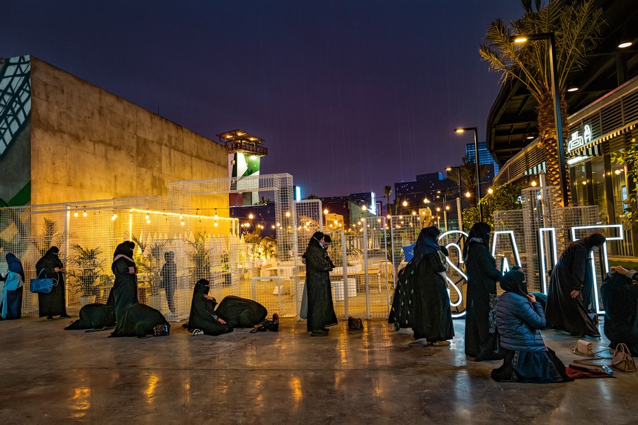 women in abayas and hijab, some standing and some praying on a polished-concrete sidewalk in front of a lit courtyard with string lights and cafe tables