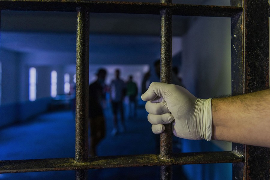 A gloved hand holds a bar on a door with several prisoners visible inside.