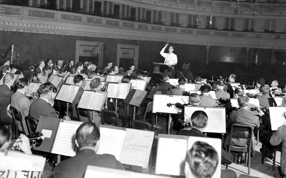 black-and-white photo of orchestra members from behind, with sheet music, on stage facing conductor with arm raised