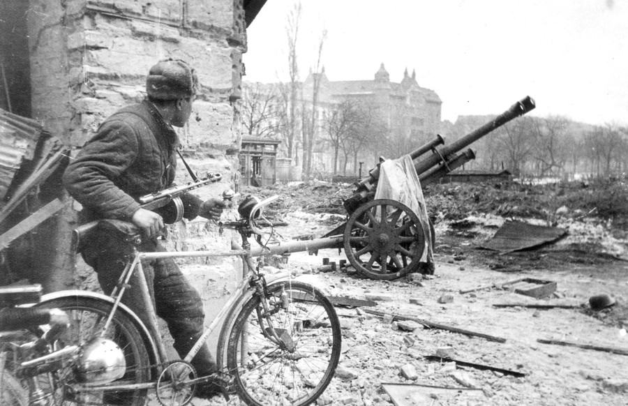 A soldier holds onto a bicycle while looking over a war-damaged city square.