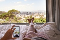 A woman takes a picture of herself lying in bed, overlooking city in the distance