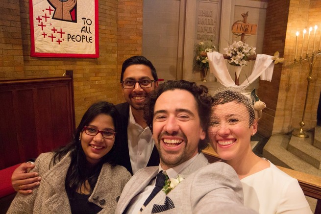 Four people smiling and hugging in a church. Abhi and his wife Abhilasha are on the left, Fernando and his wife Katherine are on the right, wearing a suit and wedding gown respectively