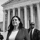 Jena Griswold, wearing a suit jacket and pearls and surrounded by men in dark sunglasses, stands in front of the Supreme Court building.