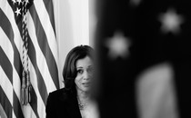 Vice President Kamala Harris is seen between two flags; one is blurred in the foreground and obscuring part of her face, and the other is just behind her.