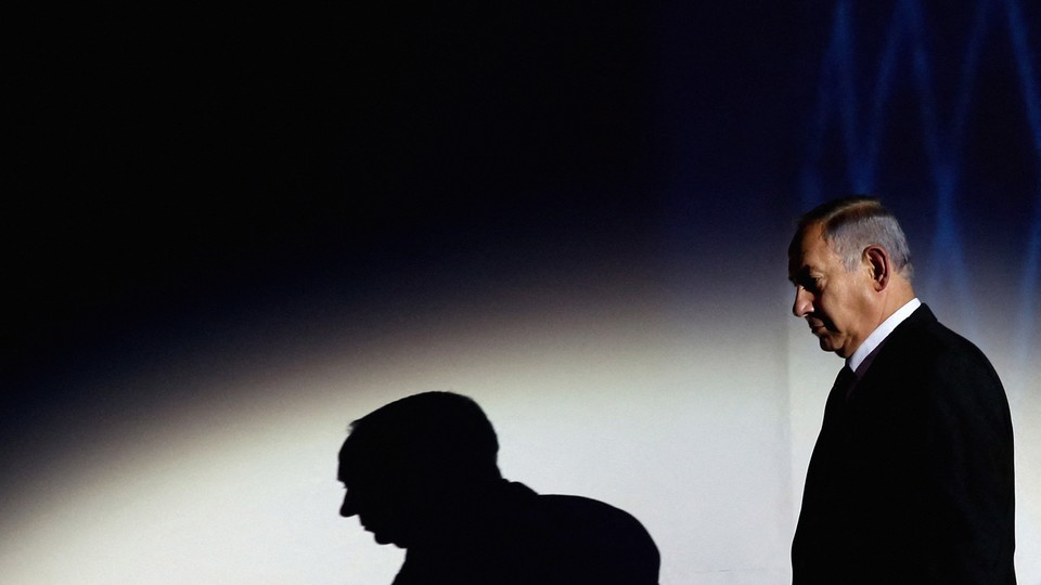 Photograph of Benjamin Netanyahu looking down with a spotlight shining on him and a shadow of himself projected on the wall behind him