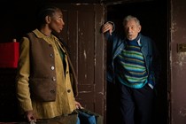 Michael Coel glances back at Ian McKellen, who stands in a doorway in an untended building.