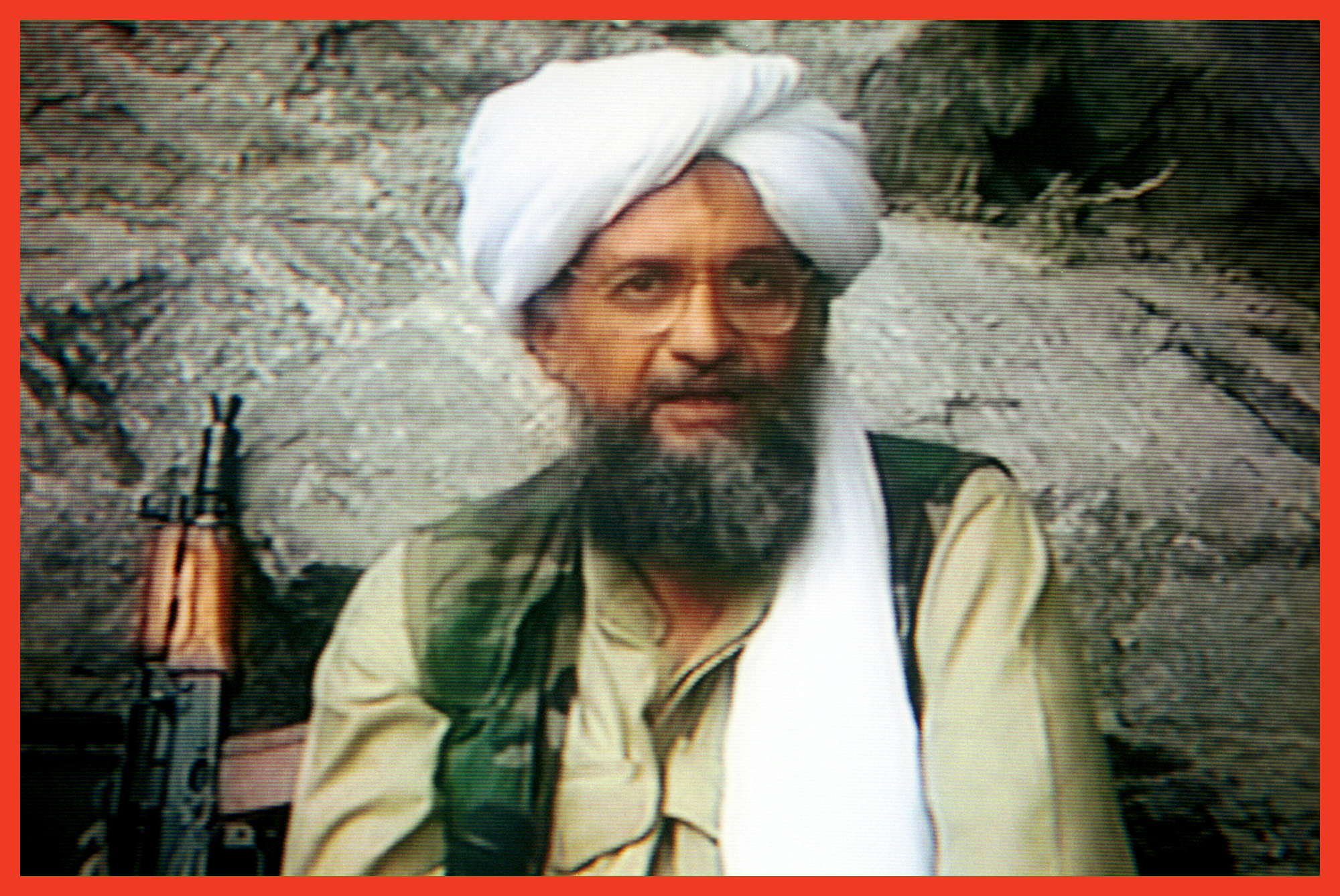 photo of bearded man in white head covering and tan jacket, wearing glasses and seated in front of rock face with rifle next to him