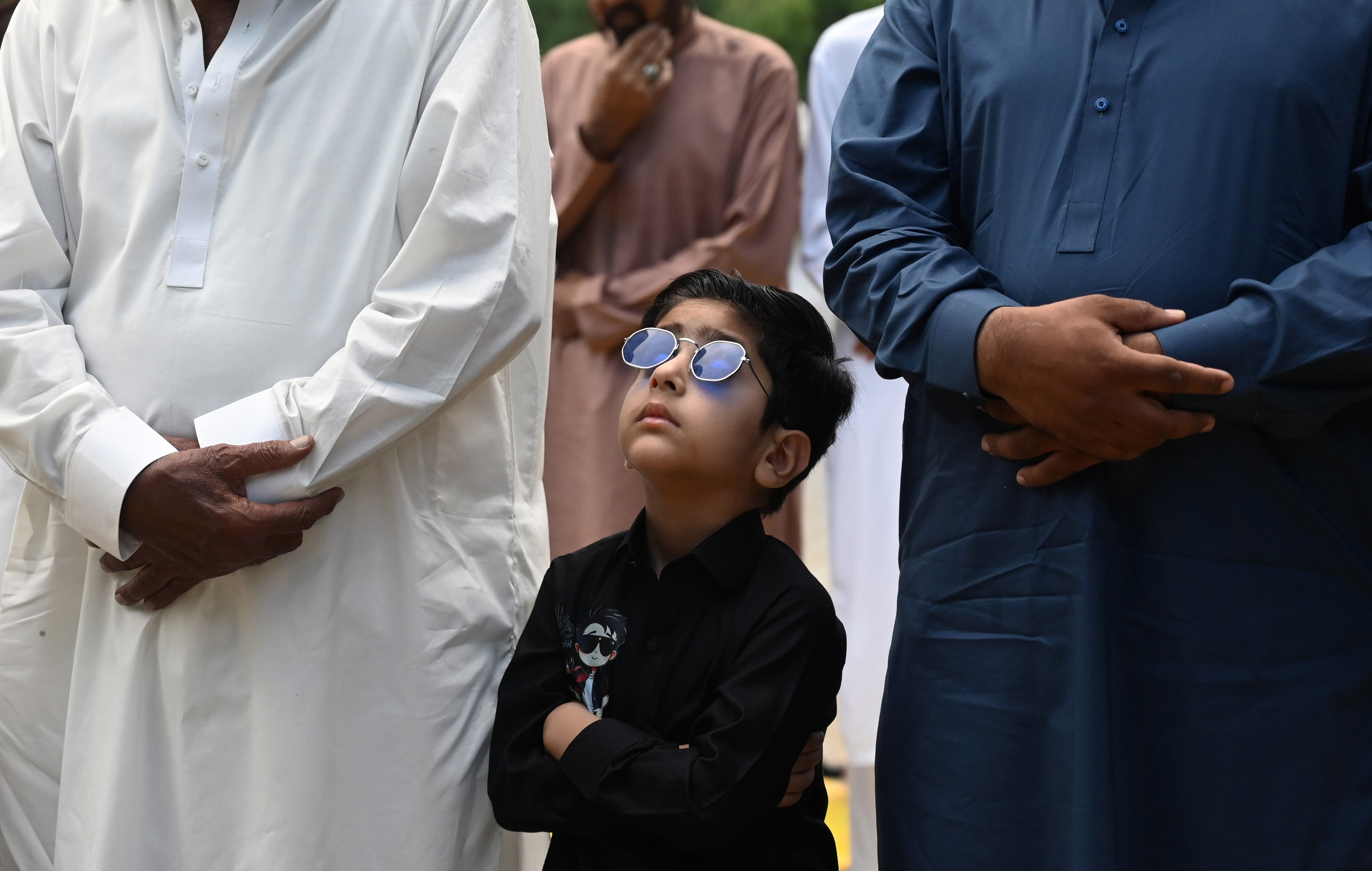 A boy wearing sunglasses looks up while standing beside adults during a Muslim prayer session.