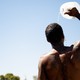 A shirtless man seen from the back standing in the sun, pouring a jug of water over his head