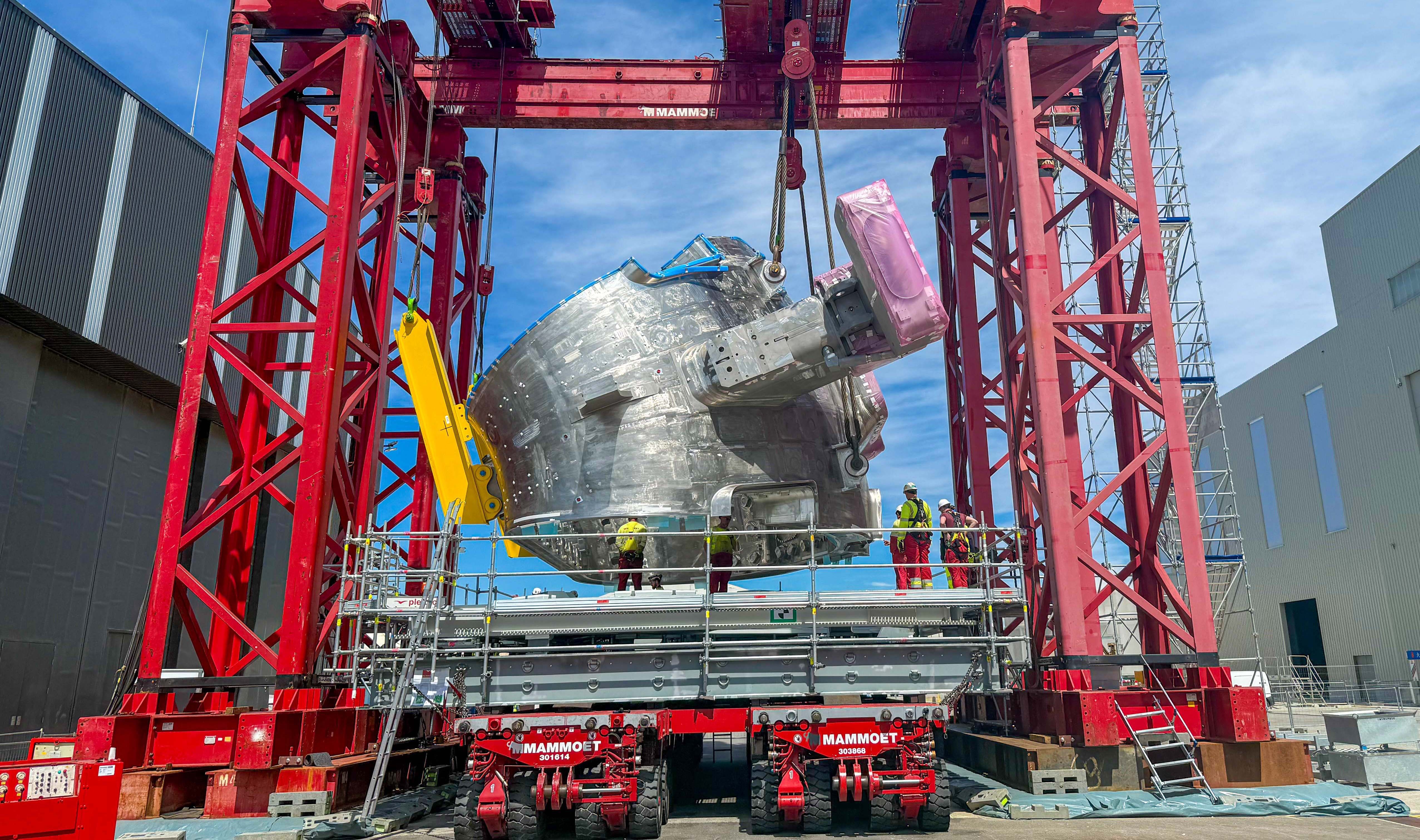 A large metal component of a fusion reactor hangs from cables in a gantry crane, while being slowly flipped.