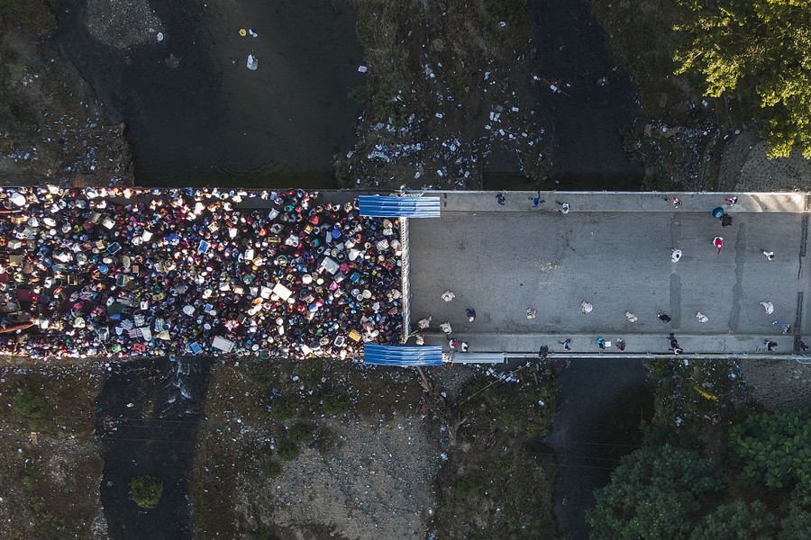 A top-down aerial view of a bridge half-filled with people waiting at a closed gate across the middle of the bridge.