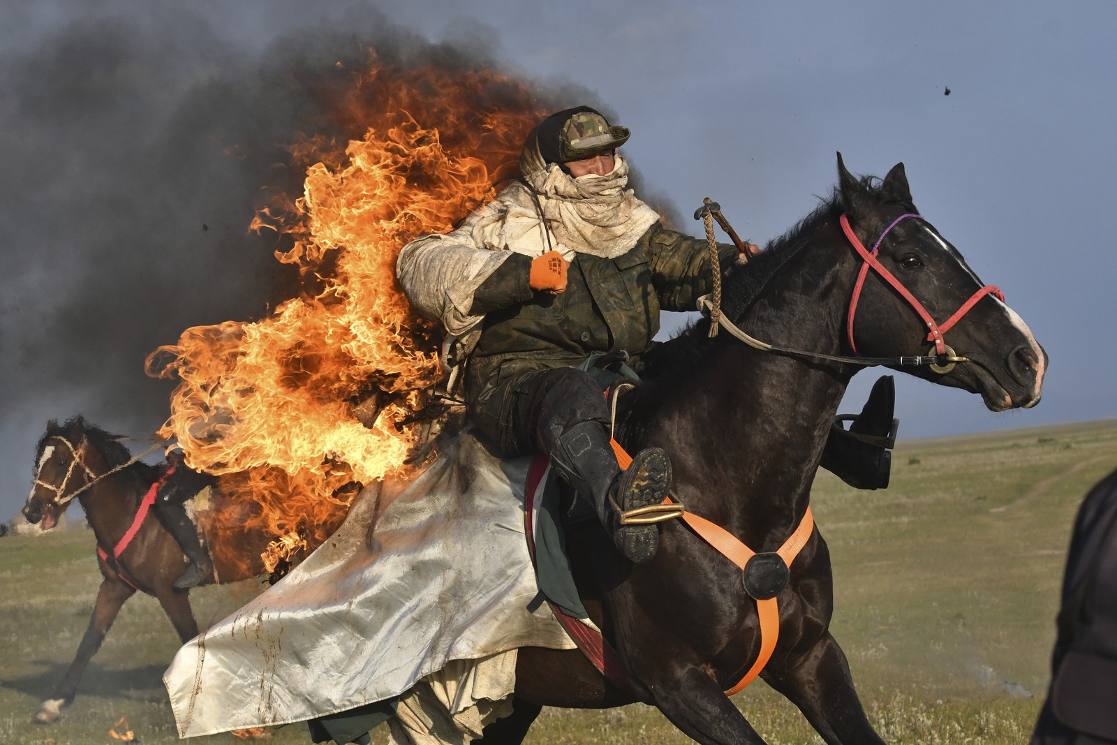 A rider on a horse, wearing flame-protective gear, rides with flames burning from their back, in a field.