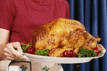 A person in a pink shirt and white skirt holding a tray with a roasted turkey on it