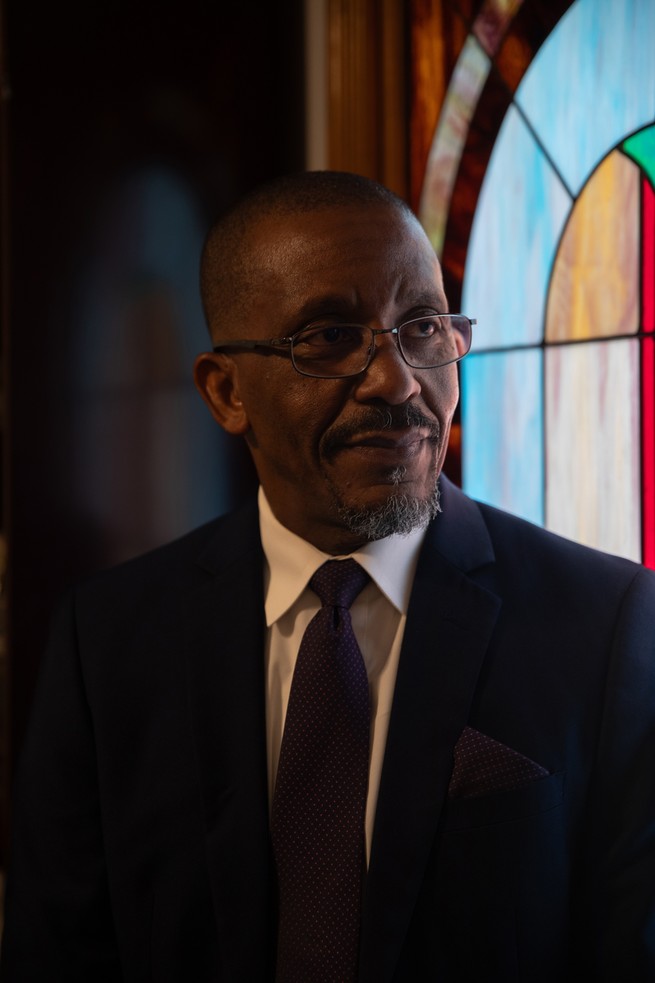 A man with glasses and wearing a suit stands for a portrait in front of stained glass. 