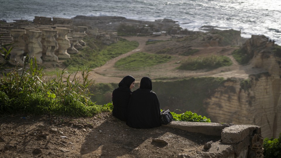 A color photograph of two women dressed in black sitting on a cliff face overlooking the water in Beirut.