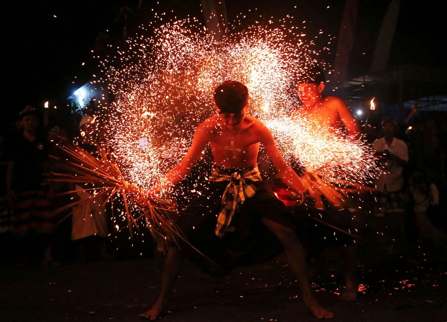 A person leans forward, reacting as others strike his bare back with burning leaves, sending up a cloud of sparks.