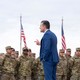A color photograph of Pete Hegseth speaking in front of uniformed soldiers on a cloudy day; four American flags are seen in the background.