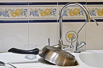 A kitchen sink with a pan, a glass, and a spoon sitting on the counter next to it.