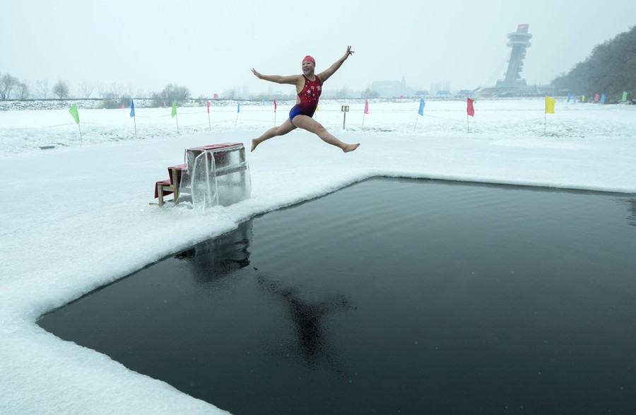 A person in a bathing suit jumps from a platform into a large opening cut into a frozen river.