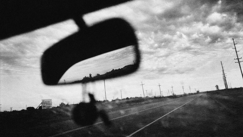 a black and white photo of the road and the cloudy sky seen out the front window of a car, the mirror taking up the front of the frame