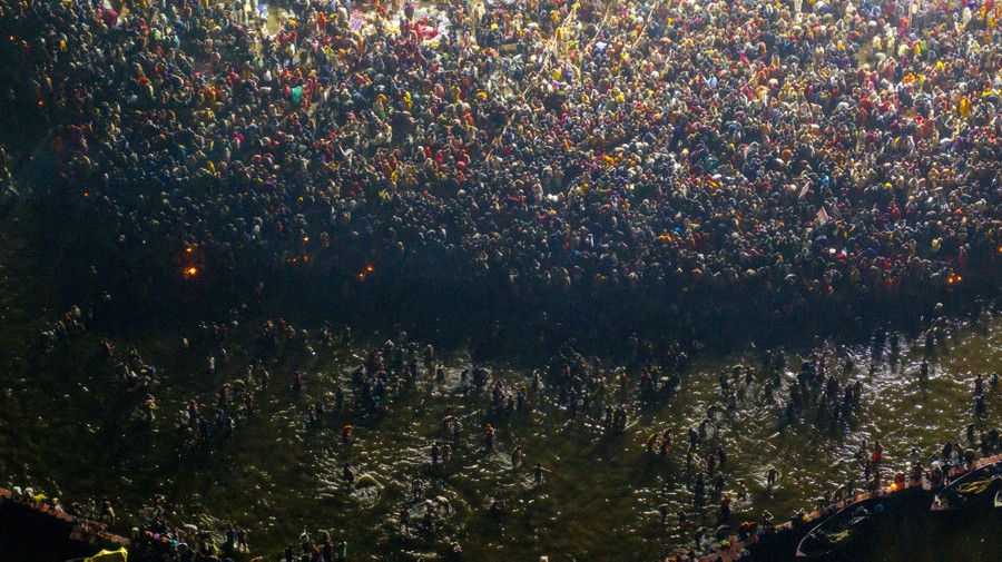 An aerial view of a large crowd along a river shore, with many others splashing and wading in the shallow water