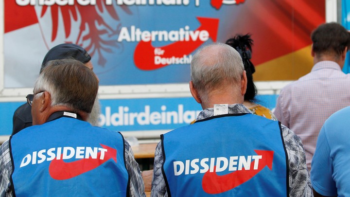 Two AfD supporters wearing the party's vests sit in front of an AfD sign.