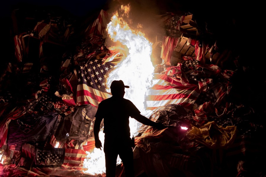 A person stands in front of a large pile of burning wooden pallets and old American flags.