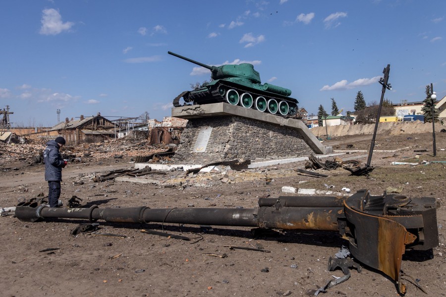 A person stands amid debris and wreckage of a tank in a square, near a World War II–era tank serving as a monument.