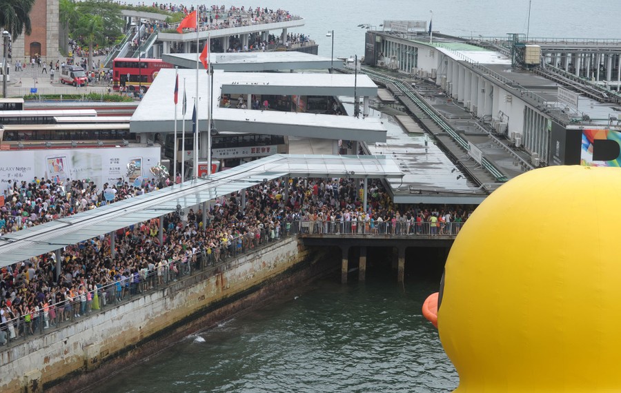 A view looks over the shoulder of a giant inflatable duck, toward crowds of people standing on docks.